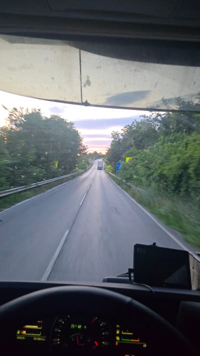The image depicts a view from the driver's seat of a truck, looking out through the windshield. The truck is on a paved road with a white line marking the center and a yellow line marking the shoulder on the left. The road is bordered by lush green trees on both sides. In the distance, a white truck is visible traveling in the same direction. The sky is a soft blend of pinks, purples, and blues, suggesting either sunrise or sunset. There are several yellow warning signs on the sides of the road. The dashboard of the truck is partially visible in the foreground, showing a steering wheel and a cluster of gauges and screens. The overall scene suggests a journey on a rural road in Dolna Verenitsa, Bulgaria, during twilight.