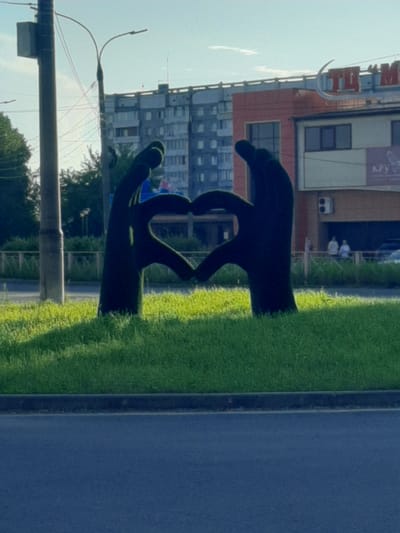 The image shows a topiary sculpture of two hands forming a heart shape. The sculpture is made of dark green material, possibly artificial grass, and is set against a background of a building and a street. The building appears to be a commercial establishment with signage. The street is paved with asphalt, and there is a grassy area next to the sculpture. The sky is visible in the background, suggesting it is daytime. The overall scene is outdoors, likely in a public space. There is no discernible activity or event taking place, as the focus is on the static sculpture. Notable details include the architecture of the building, the presence of streetlights, and the texture of the topiary. The lighting suggests it might be a sunny day. There is some text visible on the building, but it is largely illegible due to the image quality and rotation. The location is identified as Shelekhov, Russia.