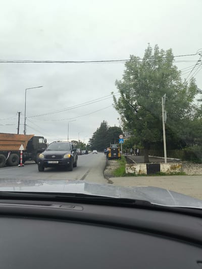 The image depicts a street scene in Martvili, Georgia, on a cloudy day. In the foreground, the dashboard of a car is visible. The middle ground features a black Toyota RAV4 with license plate TT-578-BB driving on a paved road. To the left, a large orange dump truck is parked. Beyond the vehicles, a yellow CAT construction vehicle is present, along with several people walking and standing. The environment includes trees, streetlights, and power lines overhead. A pedestrian crossing sign is visible near the construction vehicle. The road appears to be under some form of construction or repair, indicated by the presence of the dump truck and construction equipment.