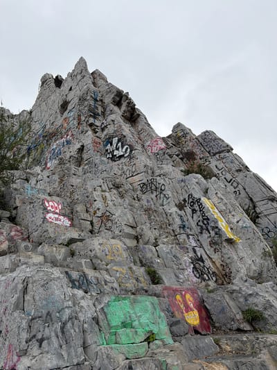 The image shows a large rock face with extensive graffiti, located in Santa Catarina, Mexico. The rock is gray with visible striations and layering. The graffiti consists of various colors, shapes, and words, covering almost the entire surface of the rock face. The sky is overcast, suggesting possible cloud cover. At the bottom of the rock formation, there is some vegetation growing. There are no visible people, interactions, or specific actions taking place in the frame.