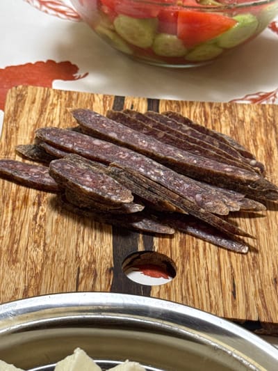 A close-up shot depicts a rustic wooden cutting board serving as the focal point, laden with thinly sliced cured sausage. The sausage slices are arranged in a haphazard pile, their color varying from dark brown to reddish-brown, speckled with lighter-colored fat particles. A glass jar containing what appears to be pickled vegetables (tomatoes and cucumbers) is visible on the left. A metal plate with pale-colored food items is located on the right side. The cutting board has a circular hole with a red-colored insert. The setting suggests a culinary scene, possibly a charcuterie board or a snack presentation. The location is unknown.
