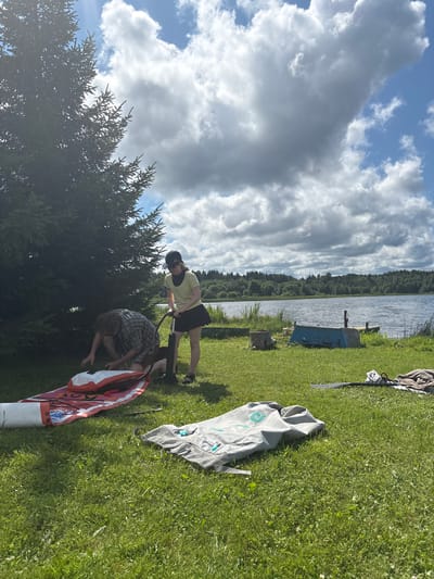 Two people are preparing an inflatable stand-up paddleboard (SUP) on a grassy area next to a lake. A young person wearing a yellow shirt and a black skirt is holding a pump, actively inflating the SUP. Next to them, another person in a plaid shirt is kneeling and appears to be assisting with the SUP, possibly holding it steady or adjusting it. The SUP is red and white with some text on it, laid out on a pink mat. In the foreground and mid-ground, other items like a folded grey bag and rolled-up towels are present on the grass. The background features a serene lake, a line of trees, and a cloudy sky. The scene is set in Chikino, Russia.
