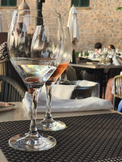 The image depicts an outdoor dining setting in Deià, Spain, likely on a terrace or patio. Two wine glasses are prominently featured in the foreground, with one containing clear liquid that refracts light into a rainbow of colors, and the other appearing to hold rosé wine. The glasses are placed on a dark, textured placemat on a table. In the background, a stone building with green shutters is visible, along with white umbrellas and other tables set for dining. blurred figures of people can be seen at tables in the distance, suggesting a restaurant or cafe environment. The lighting suggests it is late afternoon or early evening, with the warm sunlight casting reflections on the glasses and surrounding surfaces.