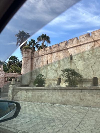 An architectural facade with faux battlements extends into a blue sky dotted with cirrus clouds. Palm trees peek over the wall. The structure features textured, aged stone and decorative corbels. The photograph, possibly taken from a moving vehicle, captures a reflection in the car window, obscuring the bottom portion of the scene. Paved ground is visible on the right.