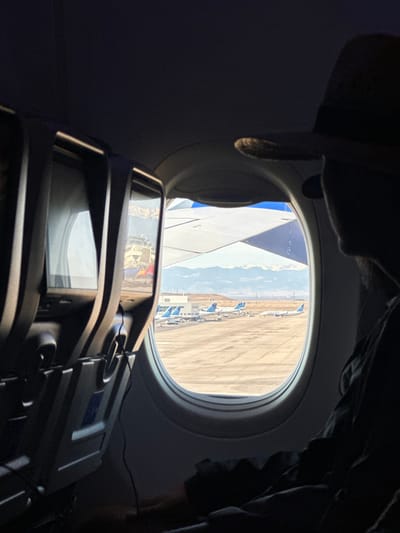 Inside an airplane, a passenger's view through the window shows a wing and an airport runway. Several parked airplanes are visible against a backdrop of mountains. An in-flight entertainment screen is mounted on the seat in front, reflecting the outside scene. The interior of the plane is dimly lit, with dark shades along the upper right and top left corners.