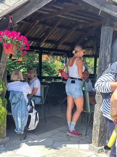 This image depicts an outdoor seating area under a wooden pergola in Dulovine, Montenegro. The foreground features a woman standing, wearing a white tank top, denim shorts, grey socks, and red sneakers. She has blonde hair pulled back and is wearing sunglasses. She is holding a red and orange bag over her shoulder. To her left, two women are seated at a table. One woman, facing away from the camera, wears a white t-shirt and has blonde hair. A denim jacket is draped over the back of her chair, next to a white and black backpack. The other seated woman, also with blonde hair, wears a white t-shirt and blue jeans, and is holding a glass up to her mouth. A bottle of water and a glass are visible on their table. In the background, lush green foliage is visible behind the pergola. The ground is paved with irregular stone slabs. A person wearing a striped shirt and dark pants is partially visible on the right side of the frame, holding a yellow pouch.