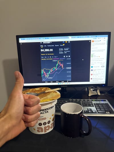 A close-up, eye-level shot captures a person's left hand giving a thumbs-up gesture in front of a computer monitor. The monitor displays a cryptocurrency trading interface showing the BTC/USDT price at 64,386.00, with a candlestick chart and associated data. To the right of the monitor, a social media feed is visible, featuring a post from "Bitcoin professor" about a giveaway. In the foreground, a container of peanut paste and two crepes with a spoon are placed on a white plate. A dark-colored mug sits on the desk to the right of the peanut paste. The background is a plain wall. The scene appears to be in an indoor setting, possibly a home or office, with no specific city or location identified.