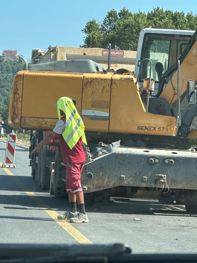 An individual wearing a red shirt, red shorts, black socks, and grey athletic shoes is standing on a paved road, partially obscured by a yellow construction vehicle. The individual has a bright yellow high-visibility vest draped over their head and shoulders, covering their face. Their right arm is extended outwards, with their hand open, palm facing away from their body. A white and red striped construction barrier is visible to the left of the individual. The construction vehicle appears to be an excavator or similar heavy machinery, with "GENEX 578" visible on its side. The road has yellow lane markings. In the background, there are trees, a building on a hillside, and a clear blue sky. The scene appears to be a road construction or maintenance area in Prijeradi, Montenegro.
