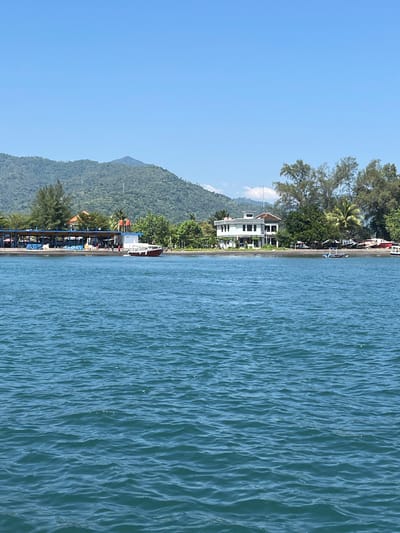 The image shows a coastal view in Pemenang Timur, Indonesia. The left side of the frame features a lush green hillside with scattered buildings and vegetation extending along the shoreline. A few structures, including a larger white building, are visible among the trees. The right side of the image depicts the ocean with visible wave patterns and a blue hue, reflecting the clear sky above. The coastline is sparsely populated with boats and structures, suggesting a relatively undeveloped or sparsely populated area.