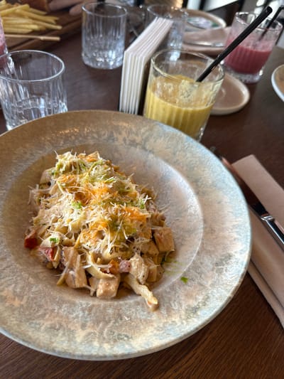 The image displays a dining scene on a dark brown wooden table, featuring a main dish and multiple beverages. Centrally positioned is a round, textured ceramic plate, primarily off-white with grey and beige speckles, holding a portion of pasta. The pasta, likely tagliatelle or fettuccine, is mixed with cubed light-colored meat (possibly chicken) and diced red vegetables, coated in a creamy sauce. It is generously topped with grated white cheese, shredded green garnish (possibly herbs), and scattered orange fragments (potentially grated carrot or spice).
To the right of the pasta, a clear glass contains a vibrant yellow smoothie or juice with a black straw. Further back and to the right, another clear glass holds a reddish-pink smoothie or drink, also with a black straw. On the left side of the table, a clear, empty glass with vertical ridges is visible. In the upper left quadrant, part of a wooden board holding a pile of golden french fries is present. A stack of white paper napkins in a holder is positioned behind the yellow drink. To the right of the plate, a metallic knife rests on a folded, light-colored fabric napkin. The background shows blurred elements consistent with a restaurant or cafe setting. The scene is located in Усады, Russia.