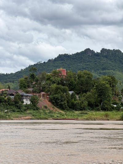 In Siao, Laos, a panoramic view captures a tranquil scene dominated by the Mekong River and lush green mountains under a cloudy sky. The foreground is occupied by the wide, muddy waters of the Mekong, with gentle ripples distorting the reflections of the overcast sky. Along the riverbank, dense vegetation and tall grasses line the shore. A narrow, reddish-brown boat is partially visible, partially submerged near the bank on the left side of the frame.
Further inland, a small village is nestled amongst the trees and hills. Several houses with tiled roofs are visible, some appearing to be under construction with wooden scaffolding. One prominent building, potentially a temple or a significant structure, features a bright red roof with ornate golden trim. The village is surrounded by a vibrant green landscape, with trees of varying sizes and shades of green dotting the hillside.
In the background, a majestic, densely forested mountain range rises steeply, its peaks softened by the cloudy atmosphere. The mountains are covered in a thick canopy of trees, with some rocky outcrops visible near the summit. The overall impression is one of peaceful coexistence between the natural landscape and the human settlement.