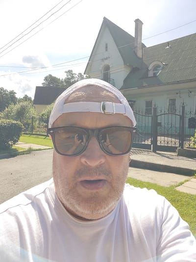 A selfie is taken by a man wearing sunglasses, a white baseball cap, and a white t-shirt. He has stubble on his face and his mouth is slightly open. The man is standing outdoors in front of a light blue house with a dark green roof and a decorative black metal gate. There is a paved road and a grassy area with trees in the background. The sky is bright and sunny with some clouds. There are power lines visible in the upper part of the image. The location is the city of Snezhinsk, Russia.