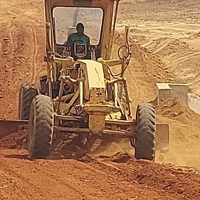 A large, yellow piece of heavy construction equipment, possibly a motor grader or a large wheel loader, is depicted in an active excavation or construction site. The image is oriented with the machine positioned diagonally, appearing to be within or alongside a trench or a steep bank of reddish-brown earth.
The machine itself is predominantly yellow, with visible wear and tear consistent with its heavy-duty use. It features large, dark, heavily-treaded tires, two of which are prominently visible on what appears to be the machine's right side, hugging a steep dirt bank. Another tire is partially visible on the opposite side, towards the lower left.
An operator is seated inside the machine's cab, which is located towards the left side of the frame. The operator, visible from the chest up through a large rectangular window, is wearing a dark green or blue long-sleeved shirt. Their posture suggests they are at the controls, engaged in operating the machinery. Inside the cab, various levers, controls, and dashboard elements are faintly discernible.
The surrounding environment consists entirely of disturbed, reddish-brown earth, characteristic of a digging or grading operation. There's a distinct, steep bank of earth immediately to the right of the machine, while the background reveals more uneven, excavated terrain and piles of soil. A large, rectangular structure, possibly a part of the machine's blade or another piece of equipment, is partially visible in the upper right background, nestled amongst the dirt mounds.
The lighting suggests it is daytime, with a warm, somewhat soft illumination that hints at either early morning or late afternoon. There are no clear shadows indicating harsh midday sun. The image quality is somewhat soft, with a noticeable grain or pixelation when viewed closely, which might be typical for a personal memory capture. No discernible text is visible on the machine or in the environment.