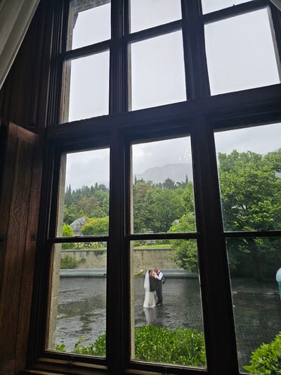 From inside a dark room, a large wooden window with multiple panes frames a view of a rainy day outside. The window’s wood is dark and ornate, with textured panels below.
Through the window, a landscape is visible. Lush green trees and vegetation dominate the scene. In one window pane, two people, a bride and groom, are visible. The groom is dressed in dark attire, and the bride is wearing a light-colored wedding dress. They stand closely together.
In the distance, a faint outline of a mountain is visible through the mist and trees. A water feature, possibly a pond or a lake, is present. Rain is falling.
The city is Алупкинский городской совет, Ukraine.