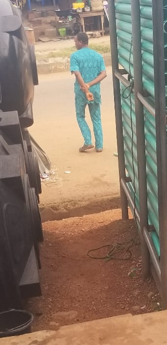 The image captures an outdoor urban scene in Ado-Ekiti, Nigeria, characterized by commercial activity and mixed ground surfaces under daylight.

In the immediate foreground, a light brown concrete surface, possibly a step, transitions to a reddish-brown dirt and gravel ground. On the left, a stack of multiple large, black, cylindrical plastic water tanks partially frames the scene, with a black bucket with a metallic handle visible on the concrete step near them. A tangled white cable and a coiled dark green rope lie on the dirt ground. A narrow dirt ditch or gutter separates this unpaved area from a light grey paved road.

On this paved road, a single adult male stands with his back to the camera, facing away. He is dressed in a light blue/teal patterned short-sleeved top and matching trousers, indicative of traditional Nigerian attire, and wears brown sandals. His hands are clasped behind his back.

In the background, beyond the man and the road, several items suggest commercial or food preparation activity. These include a wooden table supporting multiple metallic cooking pots and bowls, a black pot on a stand, and a green plastic bucket on the ground. The far background shows reddish-brown earth formations and sparse green vegetation along the road's edge.