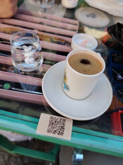 The image captures a close-up, eye-level view of a table setting, likely at a cafe or market stall in Eskisehir, Turkey. The main subjects are a small white ceramic cup of Turkish coffee, rich with dark foam, sitting on a matching saucer. Beside the coffee is a clear glass of water, which contains a small, white, stylized bunny figurine at its base.
In the blurred background, several other items are visible. To the left and behind the coffee cup, there is a small white bowl holding colorful, pastel-hued sweets, resembling Turkish delight. Further back, various pieces of jewelry, including necklaces with different stones (one green, one purple), are displayed on pink-lined trays and in blue compartments. A person's arm is slightly visible in the top-left corner, suggesting a customer or server. Part of a white dish with a blue design and a black bag are also discernible in the background.
The setting appears to be outdoors or semi-outdoors, possibly a sidewalk cafe or a stall in a marketplace, given the casual arrangement and the display of goods. The table itself is made of glass with a green frame underneath. A small, rectangular card bearing a QR code is placed on the table in the lower-left foreground, likely for payment or information. The natural lighting suggests it is daytime. The overall scene depicts a moment of relaxation, enjoying traditional Turkish coffee with its customary accompaniment of water and sweets, amidst a commercial or cultural environment.