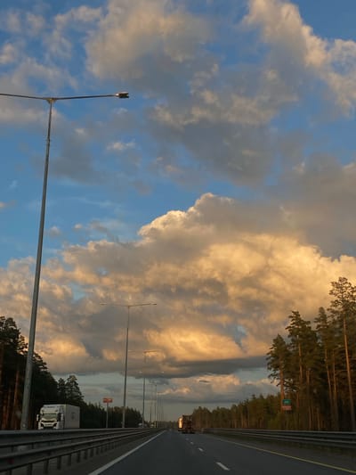 The image depicts a highway scene in Tveretskii, Russia, during what appears to be sunset. The sky is filled with dramatic, billowing clouds, tinged with warm, golden hues from the setting sun, contrasting with patches of blue sky. A prominent lamppost stands on the left side of the image, with its light turned off.

The highway itself is empty of passenger vehicles but features several trucks. On the left side of the highway, a white truck with a cargo trailer is visible. Further down the highway, more trucks are seen in the distance. The roadside is lined with trees, primarily tall pines, their silhouettes dark against the bright sky. Metal guardrails separate the lanes of the highway. Road signs are present, including a green sign with the number "265" and a red sign with white lettering. The road surface is asphalt, marked with white lines. The overall impression is one of a quiet, expansive highway under a dramatic sky.