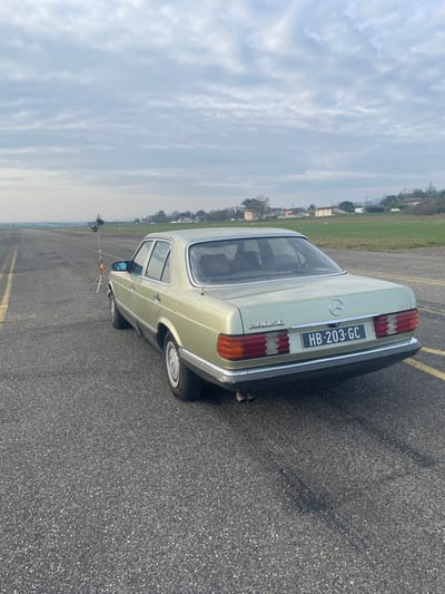 A light green Mercedes-Benz 280 SE sedan, bearing French license plate "HB-203-GC," is positioned stationary on an asphalt runway, viewed from its rear-left quarter. The vehicle's "280 SE" badging is visible on the trunk lid, and dual exhaust tips protrude beneath the rear bumper. The runway surface is coarse, dark gray asphalt, marked with faded white or yellow lines on the left. Approximately 15 meters in front of the car, on the runway's left side, a light stand equipped with a lighting fixture or reflector is present. To the right of the runway lies a flat, green grassy field. In the mid-distance, across the field, several residential buildings with light-colored facades and reddish-brown roofs are visible, backed by distant hills or mountains on the horizon. The sky is uniformly overcast. This scene is situated on an airfield, specifically the Aérodrome de Vienne Reventin, located near Chonas-l'Amballan, France. No individuals are visible. The arrangement of the vehicle and lighting equipment suggests a staged photographic or filming setup.