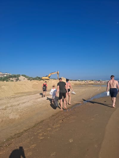 The image depicts a sunny day at a beach in Krasnaya Skala, Russia. The sky is a clear, vibrant blue, with a visible moon. Several people are present, scattered across the sandy shore. In the foreground, a group of individuals is walking along the water's edge. One man, in the upper portion of the frame, is walking away from the camera, wearing dark shorts. Further down the beach, another group of people are standing or walking together, some in swimwear.

A prominent object in the scene is a yellow excavator positioned on a sandy incline. It appears to be engaged in some form of construction or land-moving activity. In the distance, a line of buildings and more beachgoers can be seen, suggesting a populated area. The sand is wet near the water, with footprints visible, indicating recent activity. The overall mood appears relaxed and typical of a summer day at the beach. The time of day is clearly daytime, given the bright sunlight and clear sky. There is no visible text in the image.