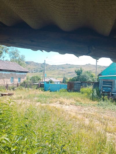 The image shows a rural scene in the city of Izgutty Ajtykov, Kazakhstan. In the foreground, there is tall, overgrown grass and some green plants. In the midground, there are several buildings and structures. On the left, a weathered, light-colored house with a tiled roof is visible, with two windows and a fence in front of it. To the right of this house, there is a blue fence. Further to the right, a rusty metal shed and another wooden shed are situated. A turquoise-colored building with a window frame is on the far right. In the background, rolling hills with sparse vegetation and a clear blue sky are visible. A utility pole with wires runs through the scene. The overall impression is one of a rustic, somewhat unkempt, rural environment.