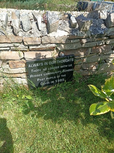 The image shows a black memorial plaque embedded in the ground in front of a dry stone wall. The plaque is inscribed with white lettering and reads: "ALWAYS IN OUR THOUGHTS THOSE NO LONGER WITH US ROSSES COMMUNITY SCHOOL PAST PUPILS OF THE CLASS OF 1985". The plaque is situated on a grassy area, with some weeds and plants growing around it. In the background, the stone wall appears to be part of an outdoor setting, possibly a graveyard or a memorial garden. The overall scene is peaceful and commemorative. The Rosses Community School is located in the town of Dungloe, County Donegal, Ireland.