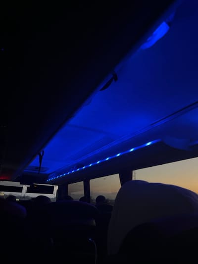 The image shows the interior of a vehicle, likely a van or bus, illuminated by blue LED lights running along the ceiling. The vehicle is likely moving, as the blurry scene outside the windows suggests. The image is taken from the perspective of a passenger seated in the back of the vehicle, looking towards the front. The passenger is in the dark, their head and shoulders obscured. The vehicle’s interior is dark, with only the blue LED lights illuminating the ceiling. The windows are tinted, and the scene outside is blurry and difficult to discern. The image appears to have been taken in Estrées-Deniécourt, France, at the coordinates 49.884° N, 2.838° E. The scene is quiet, peaceful and appears to be a simple interior shot.