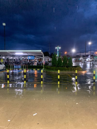 The image depicts a parking lot in Mae Chan District, Thailand, during a rainstorm at dusk. The sky is a deep, stormy blue with visible raindrops. The parking lot is flooded, with water reflecting the lights from streetlamps and a covered motorcycle parking area. The covered area, located on the left side of the frame, houses numerous motorcycles. Several black and yellow bollards line the edge of a flooded section of the parking lot, some with reflections of yellow light visible. A white car is parked on the far left, partially obscured. In the background, a landscaped island with several evergreen trees is visible, illuminated by streetlights. Power lines and poles are also present in the background. The overall scene conveys a sense of a wet and dimly lit environment.