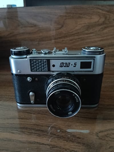 A silver and black Soviet-era Zorki-5 rangefinder camera with a Jupiter-8 50mm f/2 lens is displayed on a wooden surface. The camera is front-facing, with its lens in focus. The wooden surface appears to be a table or shelf with a subtle wood grain pattern. The background is blurred, suggesting a shallow depth of field. There is no discernible city or specific location information available from the image itself. The camera exhibits typical features of its model, including a film advance lever, a rewind knob, a shutter speed dial, and a viewfinder window. The lens has aperture and focus rings with engraved markings. The overall impression is that of a still life photograph of a vintage camera.