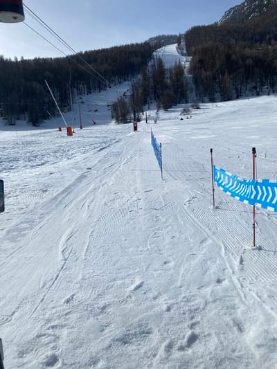 This image depicts a snow-covered ski slope in La Salle-les-Alpes, France, under a clear, sunny sky. The foreground shows groomed snow with visible corduroy patterns, interspersed with ski tracks and boot prints. On the right, a segment of blue safety netting featuring white chevron patterns is erected, supported by orange poles. A second, plain blue netting section is positioned further up the slope on the left. The mid-ground reveals a chairlift system with multiple pylons and empty chairs ascending the left side of the slope, which is bordered by dark, bare-branched trees. Orange safety markers line portions of the groomed path. A small, red structure is visible to the right of the central path. Several individuals are present on the slopes; one person is seated or lying on the snow in the mid-ground, center-right, while other smaller figures, presumably skiers, are discernible further up the main slope. The background features higher, snow-covered mountain terrain with scattered trees. Strong sunlight casts distinct shadows.