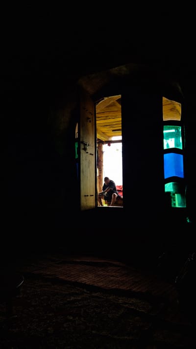 The image depicts a dimly lit interior viewed through an opening. Above the opening, there are stained glass panes in yellow, green, and blue, allowing light to filter through. The main scene is viewed through a rectangular opening in the foreground, which appears to be a wooden surface with some metal objects, possibly tools, and what look like nails or screws.

Through this opening, we see a person, likely a man, bending over. He is wearing a dark t-shirt and dark shorts. He is positioned in what appears to be an outdoor or semi-outdoor area, with wooden planks visible on the left and a bright, washed-out background suggesting daylight. The man seems to be engaged in some activity, possibly working on something or retrieving an object.

The overall impression is one of a confined, rustic space looking out into a brighter area. The lighting suggests it could be daytime, given the bright background. The texture of fabric is visible on the right side of the image, adding to the sense of an interior space. There is no visible text in the image. The location context provided is Shila, India.