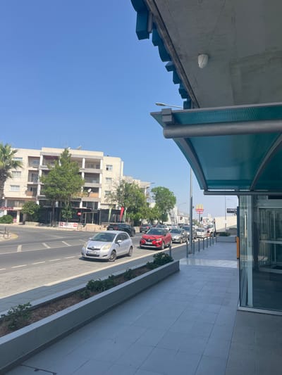 The image depicts a street scene in Latsia, Cyprus, on a clear, sunny day. In the foreground, a modern bus stop or covered walkway with a turquoise roof and concrete supports is visible. The walkway's floor is tiled with large, light-colored square tiles. A planter box with low green shrubs borders the walkway and street.
The street itself is paved and shows lane markings. Several cars are in motion or stopped in traffic. A silver Toyota Yaris, license plate MKA 545, is in the left lane, and a red Renault Clio, license plate RZT 493, is in the right lane behind it. Other vehicles are visible further down the road, including a white SUV and a black Audi.
Across the street, a multi-story apartment building with white and beige facade, balconies, and numerous windows stands. Trees and some palm trees are present in front of and around the building. Several shops are visible at the ground level of the building, with signage including "Pata" and "Illy". Further down the street, a McDonald's restaurant sign is visible, and a sign reading "CONSTA" is also present. A street lamp is visible on the left side of the image. A security camera is mounted on the underside of the bus stop structure. The sky is a clear, bright blue.