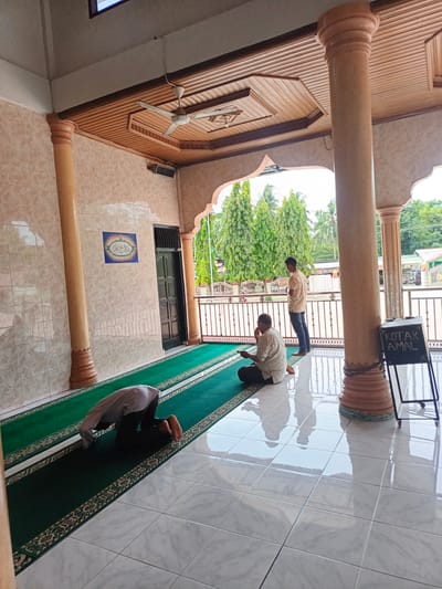 The image depicts the interior of a mosque in Sawang, Indonesia, likely the prayer area located on a porch or open-air section. Two men are present. One man is in the foreground, kneeling on a green prayer rug with his head bowed in prayer, facing away from the camera. He is wearing a light-colored shirt and dark trousers. In the mid-ground, another man stands near a railing, also facing away from the camera, wearing a tan shirt and blue jeans. Beyond the railing, lush green trees and a building are visible, suggesting an outdoor environment. The mosque features tiled walls, large cylindrical pillars, and a wooden ceiling with a fan. A blackboard with "KOTAK AMAL" (charity box) written on it is positioned near a pillar in the foreground. The floor is covered with white tiles that reflect the light.