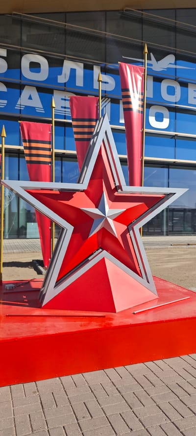 A bright red star monument sits in front of a building in Bolshoye Savino, Russia. The star is outlined in gray and sits on a red platform. Behind the star are three red flags with orange and black stripes. The building in the background has blue and white lettering that reads "БОЛЬШОЕ САВИНО". The ground is paved with gray brick.