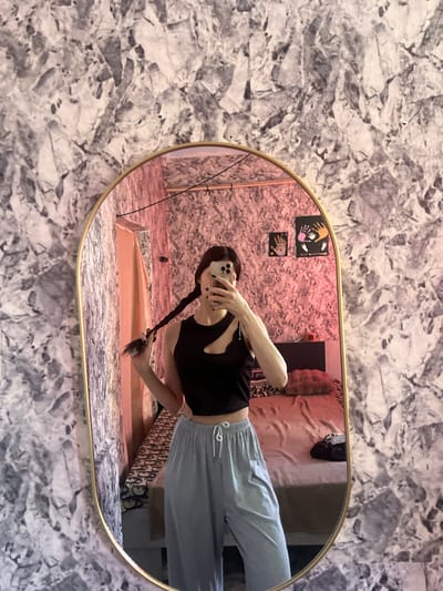 A young woman with dark hair styled in a braid poses in front of a mirror, taking a selfie. She is wearing a black asymmetrical crop top with a cut-out detail and light blue lounge pants. Her left hand is holding her braid, and her right hand is holding a smartphone to take the picture. The mirror has a gold frame and is oval-shaped. The background visible in the mirror shows a bedroom with a bed covered in pink bedding, a pillow, and a dark bag on the bed. The walls in the room are covered with a patterned wallpaper. The overall setting appears to be an indoor residential space. The location is not specified in the provided image or crops.