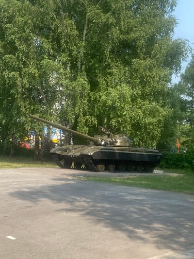 A green military tank is parked on an asphalt surface in front of a dense row of birch trees. The tank is oriented diagonally with its barrel extending to the left. The trees are full of green leaves, and the sky above is clear blue. In the background, behind the trees, there are hints of buildings and structures that appear to be part of a city. The asphalt surface in the foreground has some white chalk markings and shadows cast from the trees. This scene is located in the city of Mamontovo, Russia.