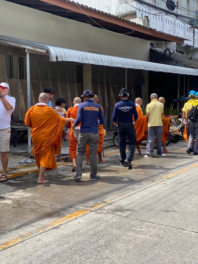 This forensic description details a daytime street scene in Ban Phe, Thailand. A procession moves along a concrete street with faded yellow lines and visible stains. Approximately four bald, barefoot Buddhist monks, draped in saffron/orange robes, are central to the image. They are engaged in interaction with civilians, consistent with an alms round.

On the far left, a light-skinned man in a white polo shirt, beige shorts, and sandals stands, observing the group with his left arm raised. Towards the center-left, behind the leading monk, civilians including a person in a patterned top are seen extending their hands towards the monks, suggesting alms giving.

Two men in dark blue long-sleeved shirts are prominent. One, in the center, wears grey trousers and a backward baseball cap, with "กรมประมง" (Department of Fisheries) in white Thai script on his back. Another man in a similar dark blue shirt with a white emblem and backward cap stands further right. These individuals appear to be officials or organizers.

Other participants include a man in a light yellow polo shirt and light grey trousers on the right, and several individuals in yellow shirts and blue caps further right, one with a black backpack.

The background consists of a row of older two-story buildings, likely shophouses, with closed ground-floor storefronts and a continuous corrugated metal awning extending above. Overhead, electrical wires are visible. A white scooter is partially visible on the right side of the street. The bright sunlight casts distinct shadows on the ground and subjects.