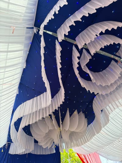 The image shows an interior view of what appears to be a decorated tent or canopy, possibly for an event or celebration. The background is a dark blue material dotted with small lights, reminiscent of a night sky. Across this backdrop are draped layers of white fabric, some arranged in wavy, ruffled patterns and others in a more linear, cascading style. The image is taken from a low angle, looking upwards toward the ceiling of the structure. The location is Tay Son Commune, Vietnam.