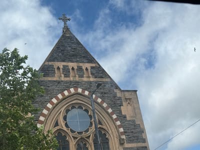 An upward-angled outdoor shot captures the facade of a historic stone building, likely a church, against a partially cloudy blue sky. The building's prominent feature is its dark grey, rough-hewn stone construction, contrasting with lighter, smooth stone accents. A large, ornate stone cross crowns the apex of a steep gable roof. Below the gable, a horizontal band of lighter, rectangular stones features several small, arched niches.
The lower part of the facade is dominated by a grand arched entrance or window opening, which frames a circular rose window with intricate stone tracery. The arch itself is distinctive, with alternating sections of light and reddish-brown stone forming a striped pattern. A tall, black street lamp pole stands directly in front of the rose window, partially obscuring its details. To the left, vibrant green tree branches and leaves extend into the frame, partially covering the building's stonework.
On the right side of the building, a buttress-like structure with a tall, narrow arched niche is visible. Two birds are seen flying in the distance against the sky; one small bird is in the upper left, and a larger bird with spread wings is in the upper right. The scene depicts a clear, cloudy day. No specific activities or events are taking place beyond the flight of the birds. There is no visible text. The architecture suggests a European, likely British, setting, consistent with the given location hint of Bristol, United Kingdom.