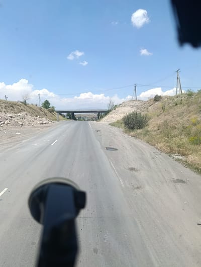 The media file shows a view from a vehicle traveling on a road in Jrvej, Armenia. The road is paved but shows signs of wear, with potholes and scattered debris. On either side of the road are dry, grassy hills and rocky embankments. In the distance, a concrete overpass spans the road, suggesting an intersection or highway. Power lines and poles are visible along the roadside, indicating infrastructure. The sky is bright blue with scattered white clouds, suggesting a clear, sunny day. The overall scene is rural and somewhat arid, typical of a summer day in this region. There is no visible text in the image. The activity appears to be a journey or drive. The foreground is partially obscured by a dark, blurry object, likely a piece of equipment within the vehicle such as a dashcam or phone mount.