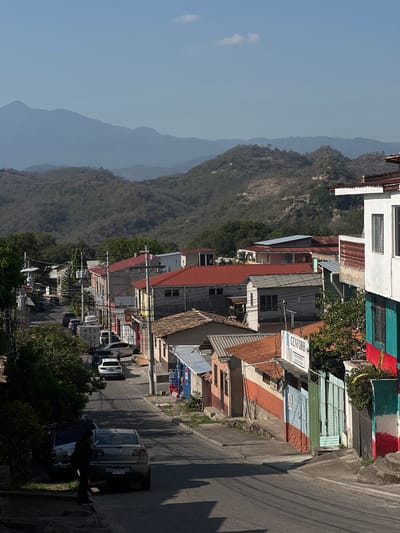 The image shows a street scene in Gracias, Honduras, with mountains visible in the background. A mix of residential and commercial buildings lines the street, some with colorful paint. Cars are parked along the street, and a person is standing next to one of the vehicles. Overhead power lines are strung between poles along the street. The mountains in the distance are covered in dense vegetation.