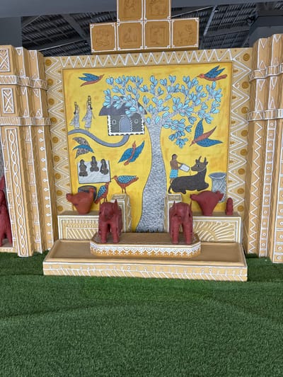 An elaborate indoor cultural display from Bhopal, India, is presented on a surface of artificial green grass. The central feature is a large, rectangular painting framed by a multi-tiered, golden-yellow structure with white geometric patterns.

The painting, set against a vibrant yellow background, depicts a stylized rural scene. A prominent, tall tree with a textured grey trunk and numerous light-blue leaves dominates the center. A small, grey house with a dark roof and white window frames is integrated into the tree on the left. On the left side of the painting, three dark-skinned women, dressed in light attire and carrying pots on their heads, walk along a curving grey path towards the tree. Below them, three dark-skinned figures are seated on the ground. On the right side of the painting, a dark-skinned male figure, wearing blue shorts, is shown feeding a dark-colored bovine with a white bowl; a white woven basket is positioned nearby. Several stylized blue and red birds are depicted flying or perched throughout the painting.

The main painting is flanked by two golden-yellow vertical pillars adorned with white geometric patterns. Above the painting, a golden-yellow horizontal structure supports a series of relief-carved square panels. The top-most panel features a stylized head, and directly below it are six smaller square panels, arranged in two rows of three, each depicting various human and animal figures in relief.

In the foreground, on a multi-tiered golden-yellow platform with white decorative trim, several reddish-brown terracotta figures are arranged. Two stylized elephant figures stand on the slightly raised central section of the platform. On the lower platform, to the left, is a stylized terracotta bird figure, and to the right, another similar bird figure along with a small, upright, cylindrical terracotta object. The entire display is viewed from a frontal perspective, with a grey ceiling structure visible above.