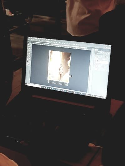 A dark-colored laptop is depicted in a dimly lit setting in Panhauya, Nigeria. Its screen displays a photo editing application, likely Adobe Photoshop, centered on a vertical image of a light-skinned male. The male subject stands on steps, dressed in a light-toned garment, illuminated by warm light, with balloons visible in the background. The software interface shows toolbars on the left and right. Partially visible hands operate the laptop's keyboard and trackpad. The background is largely obscured by shadow, with a light-colored, amorphous object visible in the upper-center.