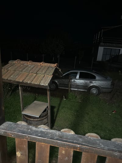 The image shows an outdoor scene at night in what appears to be a backyard or garden setting. A silver car is partially visible behind a chain-link fence. Adjacent to the fence is a wooden well structure featuring a barrel and a tiled roof. The scene is possibly located in the city of Drachevo, Bulgaria. The image is taken from an elevated position, overlooking the area. The lighting is dim, with the ambient light primarily illuminating the car and the immediate surroundings. The foreground contains a portion of a wooden structure with circular elements, possibly a fence or a railing. There are no visible people in the frame.