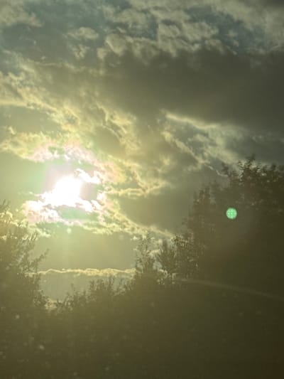 The image shows a hazy sky with the sun shining through clouds. The sun appears as a bright, white light source with a green lens flare to its right, suggesting it might be setting or is partially obscured by clouds. There are trees visible in the foreground and middle ground, silhouetted against the bright sky. The overall lighting is soft and diffused, creating a somewhat ethereal atmosphere. The environment suggests an outdoor setting, likely in a natural or semi-natural area, possibly a park or a rural landscape. There are no people or specific urban structures identifiable in the image. The location of Melivoia, Greece, is not discernible from the visual elements present in the photograph.