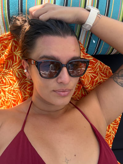 A woman is reclining, likely outdoors, wearing sunglasses and a maroon-colored top. She has fair skin, dark hair pulled back, and wears a white smartwatch on her left wrist. Her right arm is raised with a tattoo visible on her inner arm. She is lying on a striped cushion with an orange and yellow patterned pillow underneath. The location cannot be determined from the image.