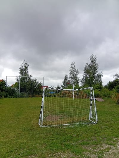 A wide-angle, full outdoor shot captures a cloudy, overcast day in Kiełczów, Poland, with a focus on a portable soccer goal on a grassy field. The goal, positioned in the foreground and slightly to the right, features a white frame with black and white striped uprights and crossbar, and a white net. In the middle ground, a smaller, similarly constructed soccer goal is visible further back on the left. A chain-link fence encloses a section of the field behind the goals. Several trees, including birches, are scattered across the landscape, with dense green foliage bordering the field in the background. A section of red-tiled roof, suggesting residential buildings, peeks through the trees on the far left. The ground is covered in green grass with some patches of dirt and gravel. The sky is uniformly gray and filled with heavy clouds. No people are present in the image.