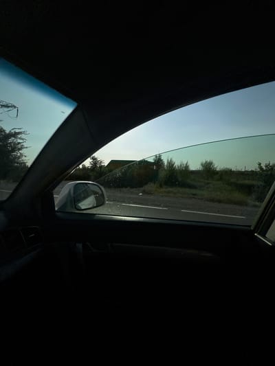 The image is taken from inside a vehicle, looking out the passenger side window. The window is partially open, with the glass angled upwards. The interior of the car, including the dashboard and door panel, is dark and mostly out of focus.
Outside the car, a paved road with a white line marking the lane is visible, running horizontally across the frame. Beyond the road, there is a grassy area with bushes and some trees. In the distance, there is a building with a dark roof. The sky is a pale blue, suggesting it is either early morning or late afternoon.
The scene appears to be a rural or semi-rural area. No people are visible in the image. The overall impression is one of a quiet, open landscape observed from a moving vehicle. The location in Istiqlol, Uzbekistan, is not discernible from the visual information provided in the image.