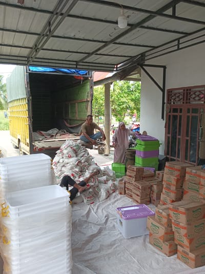 The image depicts an outdoor scene in Tanjongteungkuali, Indonesia, likely a storage or distribution area. A large, open-backed truck, partially loaded with green tarpaulin and various sacks, is parked. In front of the truck, a man in a green t-shirt and jeans sits on the edge of the truck bed, appearing to rest. A large pile of white sacks, possibly containing rice or similar goods, is stacked on the ground. Another person, wearing black pants and a dark shirt, is lying on the ground amidst the sacks, with their legs exposed.

To the right of the sacks, there are several stacks of cardboard boxes, many with visible branding such as "Sania" and "Zinc". A woman wearing a pink hijab and a long pink dress is standing near a stack of colorful plastic containers (green and purple). There are also stacks of large white plastic containers with yellow handles visible on the left side of the image. The scene is covered by a metal-framed roof with corrugated metal sheeting, providing shade. Trees and foliage are visible in the background, suggesting a somewhat rural or semi-urban environment. The ground is covered with a large white tarp or plastic sheeting. The overall impression is one of a work in progress, possibly related to the loading or unloading of goods.