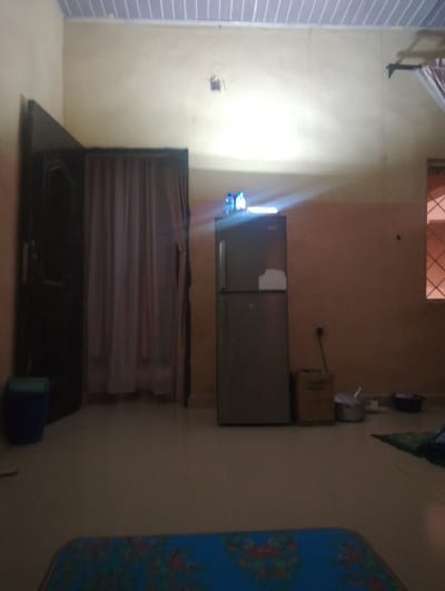 The image captures an indoor room, likely a living space or shared area, characterized by its utilitarian furnishings and lighting. The room features light-colored, possibly cream or beige, walls, with a peach or light orange wall section visible on the right side. The ceiling is made of white, horizontal slats or panels. The floor is covered with light-colored tiles.

On the far left, a dark-colored wooden door is visible, appearing to be partially ajar or set at an angle. Adjacent to the door, a light-colored, translucent curtain is drawn, obscuring what appears to be another doorway or a large window. A small blue trash can or bucket sits on the floor near the door.

Dominating the center-right of the image is a two-door refrigerator, with the freezer section on top and the main refrigeration unit below. On the very top of the refrigerator, two small plastic bottles, possibly containing water, are placed, reflecting a significant amount of light. A strong, bright light source, likely an overhead bulb or fixture that is mostly out of frame, casts a powerful glare onto the wall above the refrigerator, creating noticeable lens flare and illuminating that section of the wall intensely. A dark patch or opening is visible on the wall above the refrigerator, potentially an electrical outlet or fixture mounting. A white electrical cord runs down the wall beside the refrigerator, plugging into a socket.

Further to the right, a window with a distinctive grid pattern of horizontal and vertical bars is visible, allowing a faint amount of light to enter the room. Below the window, a brown, rectangular container or box is placed on the floor, next to two metallic pots or bowls. In the immediate foreground, a portion of a blue mat with a floral pattern is visible, suggesting a seating or sleeping area.

No specific activity is depicted, and the room appears to be in a static state of occupancy. The strong artificial light suggests the photograph was taken during the evening or night, or in a space with limited natural light. No discernible text is visible in the image.