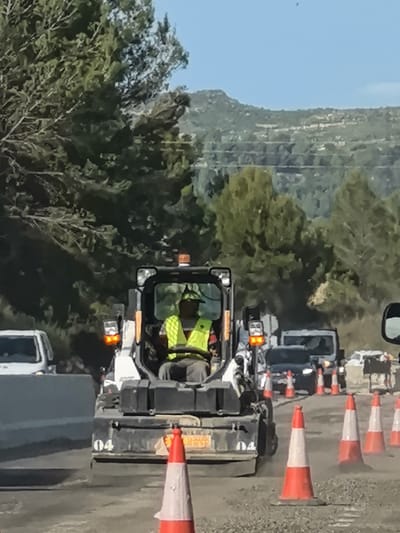 The image captures a road construction or maintenance scene under clear blue skies in a mountainous, vegetated area of Castellbell i el Vilar, Spain.
In the foreground and center, a male worker operates a compact utility vehicle, possibly a skid-steer loader, equipped with a front-mounted attachment for road work. He is wearing a high-visibility yellow vest, indicating his role in a construction environment. The vehicle has illuminated orange warning lights on its top and sides, emphasizing its presence in a work zone. The number "04" is visible on the lower front side of the vehicle's chassis, possibly a unit identification number. Faint, unreadable text, likely a license plate or branding, is also visible on the vehicle's rear panel, partially obscured, but appears to contain "CBB".
The road surface beneath the vehicle and in the immediate vicinity is composed of loose aggregate or gravel, consistent with ongoing road works. A line of orange and white traffic cones extends along the right side of the road, marking the work area and guiding traffic. On the left side, a white concrete barrier separates the work zone from what appears to be a functional road lane.
In the background, several vehicles are visible, including a white van on the left and darker vehicles further back, suggesting traffic is moving through or near the construction area. Lush green trees flank the road and densely cover the rolling hills in the distance, typical of a rural or semi-rural mountainous landscape. The bright sunlight indicates it is daytime.